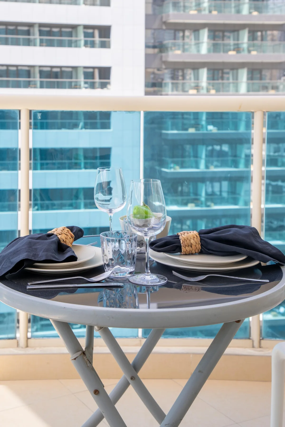 A small balcony table is set with plates, folded napkins, glasses, and a potted plant.