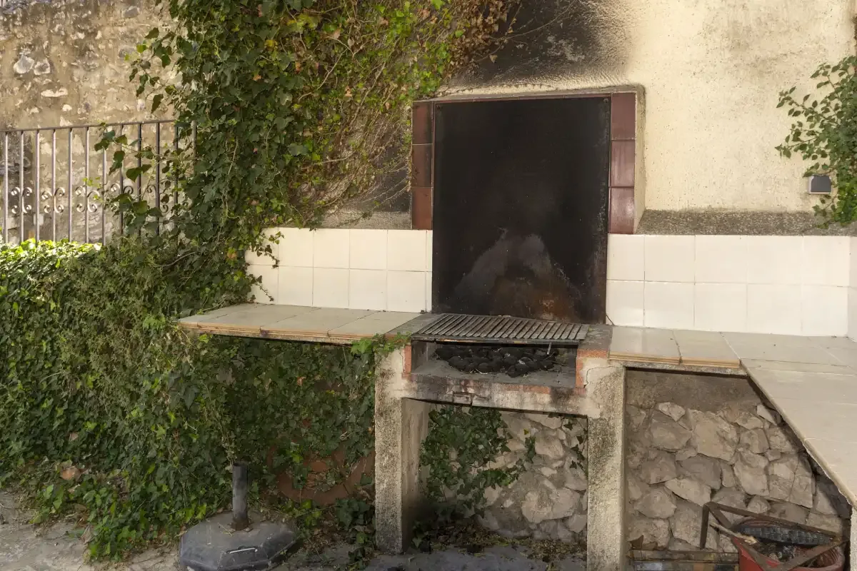 Una parrilla exterior está junto a una encimera de azulejos y plantas frondosas.