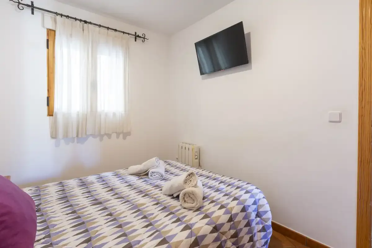 Dormitorio con ropa de cama estampada, una ventana con cortinas y un televisor en la pared.