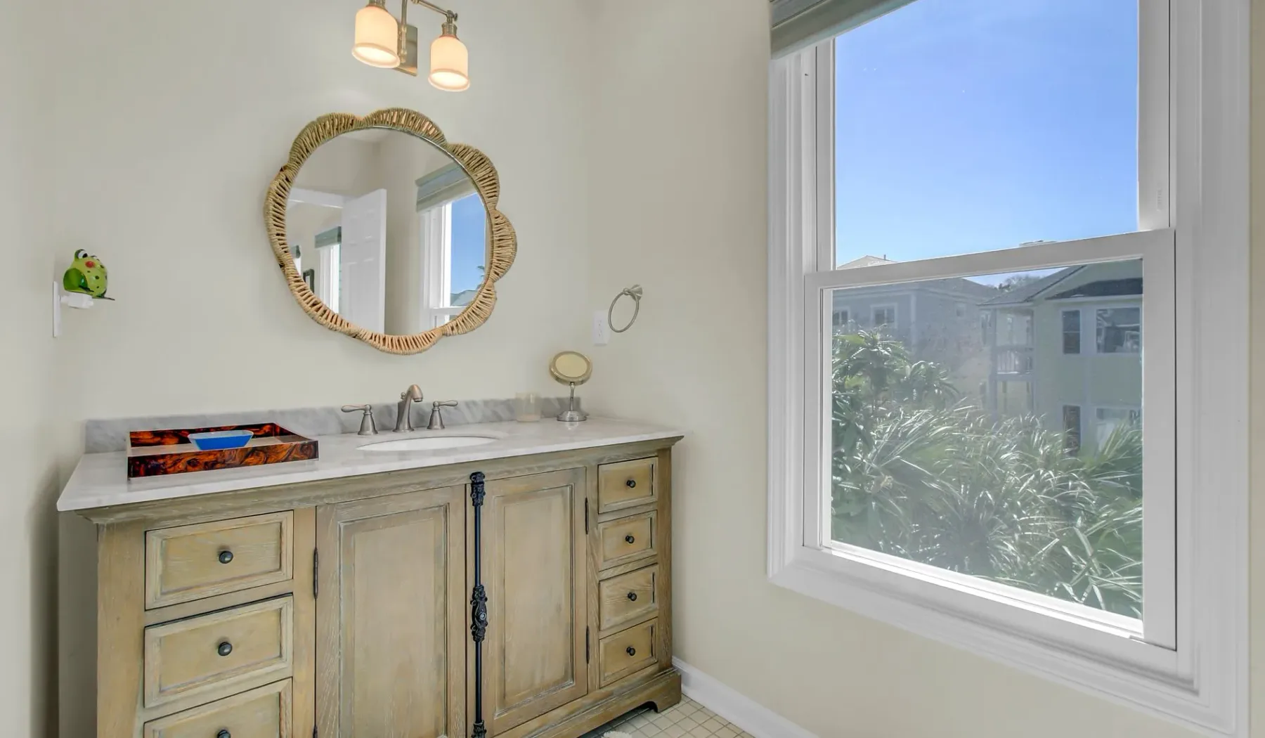 Un tocador de baño luminoso está bajo un espejo decorativo junto a una ventana grande.