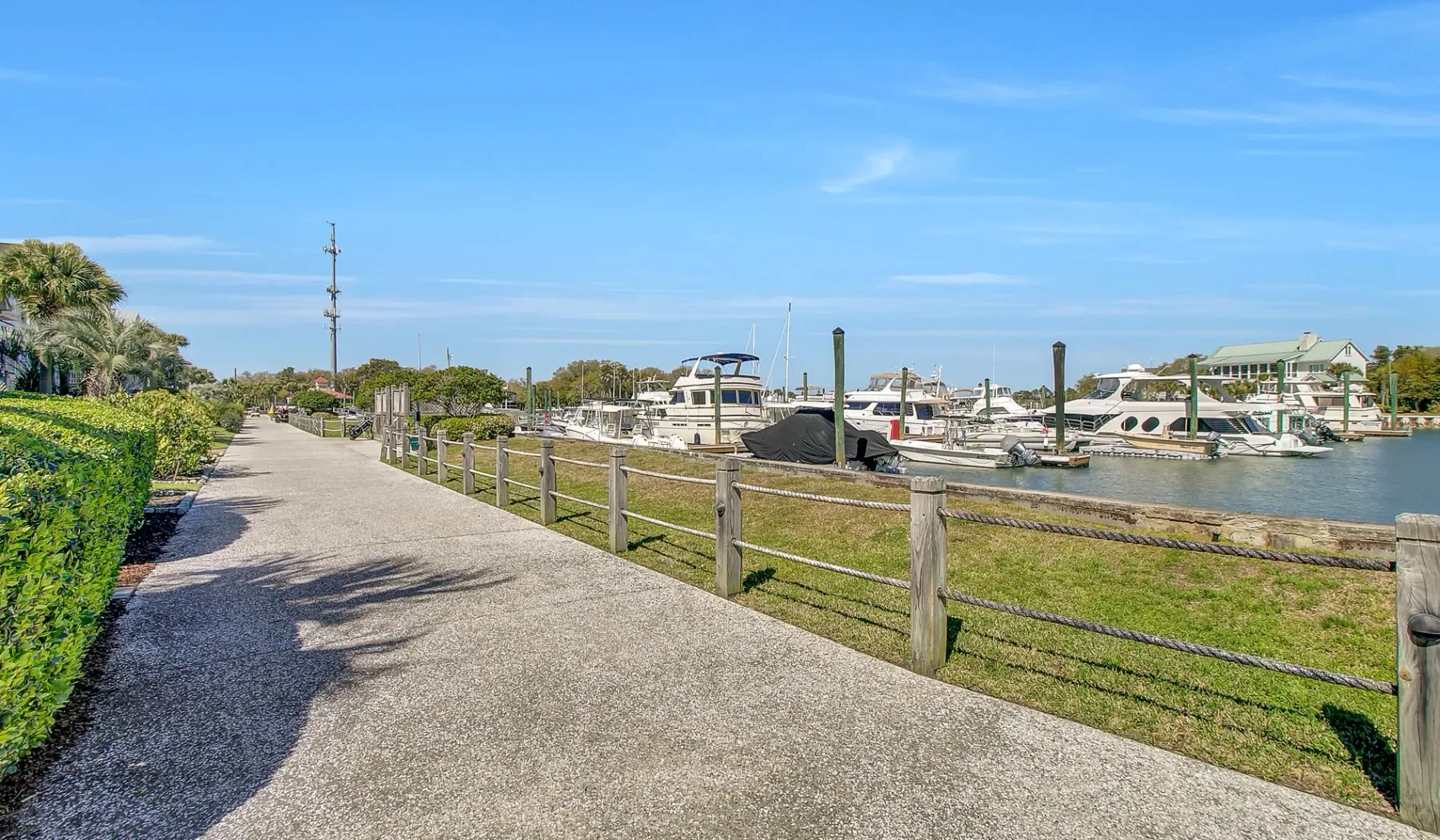 Un sendero pavimentado junto al agua bordea un puerto deportivo con barcos y vegetación.