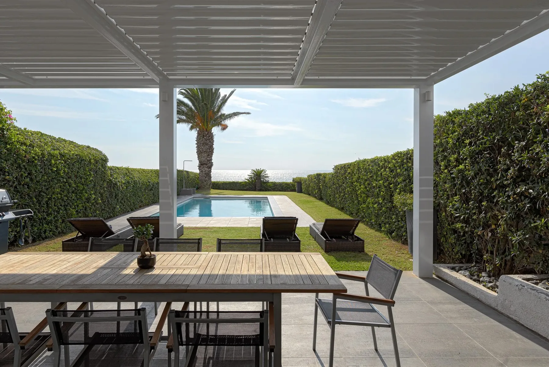 A shaded patio with outdoor seating overlooks a swimming pool and garden.