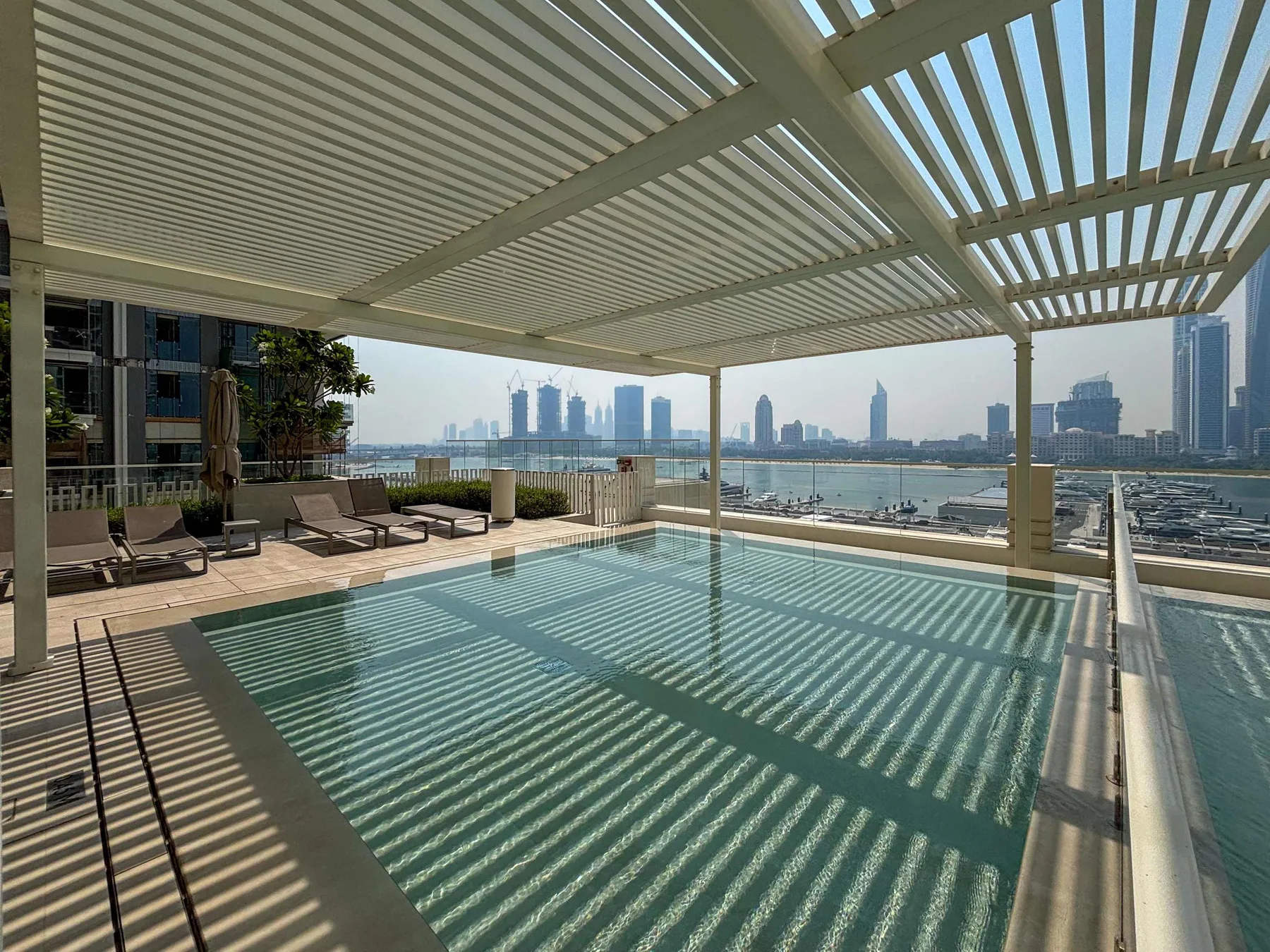 A rooftop swimming pool sits beneath a slatted canopy, overlooking the water and city skyline.