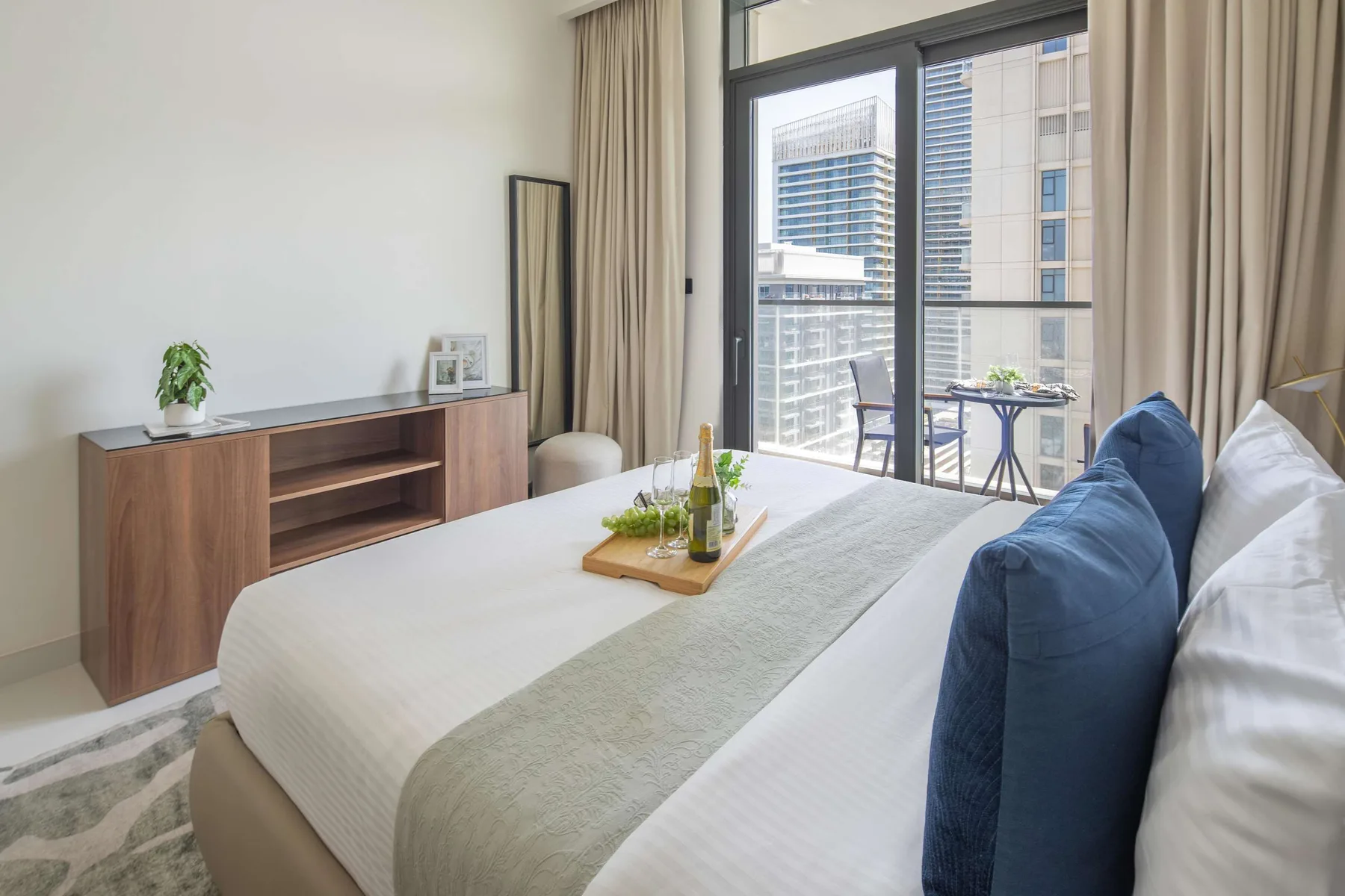 A bright bedroom with a neatly made bed and large windows overlooking nearby buildings.