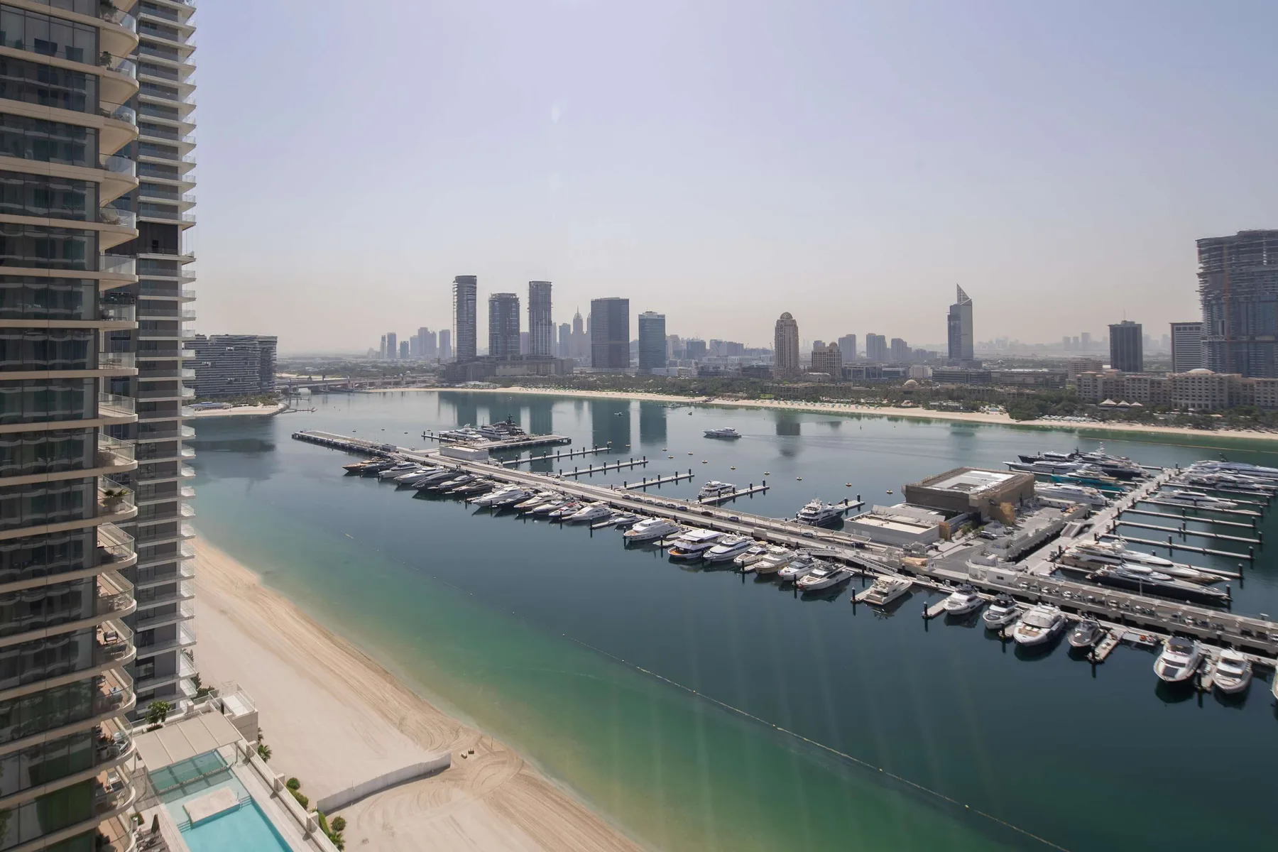 A marina with rows of boats sits beside a sandy beach and distant city skyline.