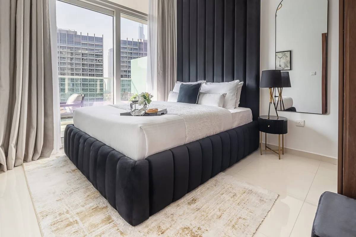 A bedroom with a bed beside large windows and a dark upholstered headboard.