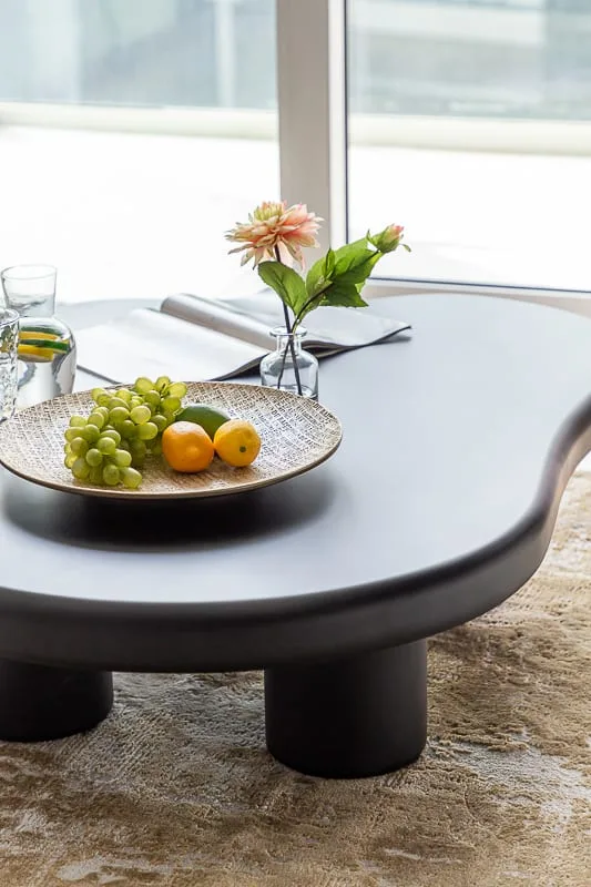 A low coffee table holds fruit, a flower vase, and a glass in a bright room.