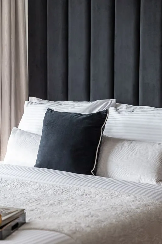 A bedroom with white bedding and a dark pillow against a tall upholstered headboard.