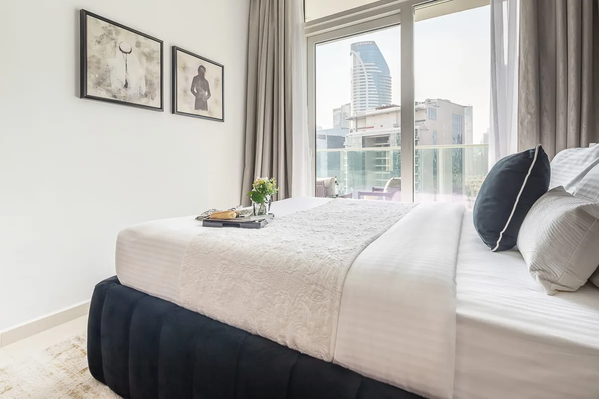 A bright bedroom with a neatly made bed beside large windows overlooking nearby buildings.