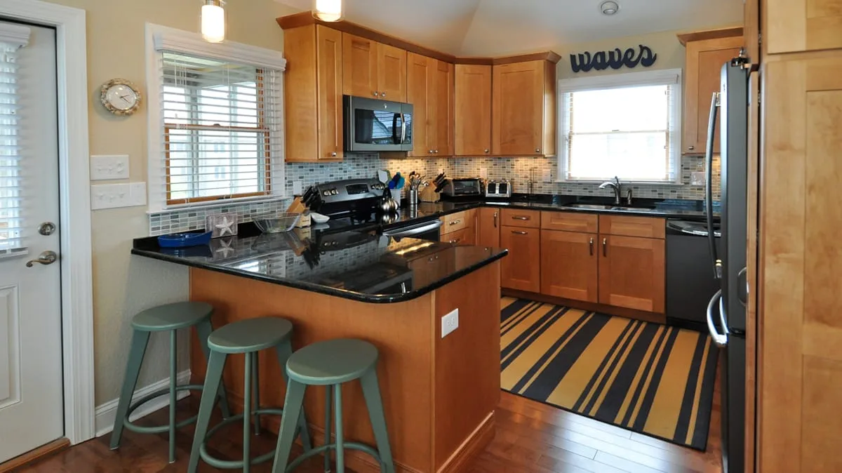 A warm-toned kitchen with wooden cabinets, dark countertops, and stools at a peninsula.