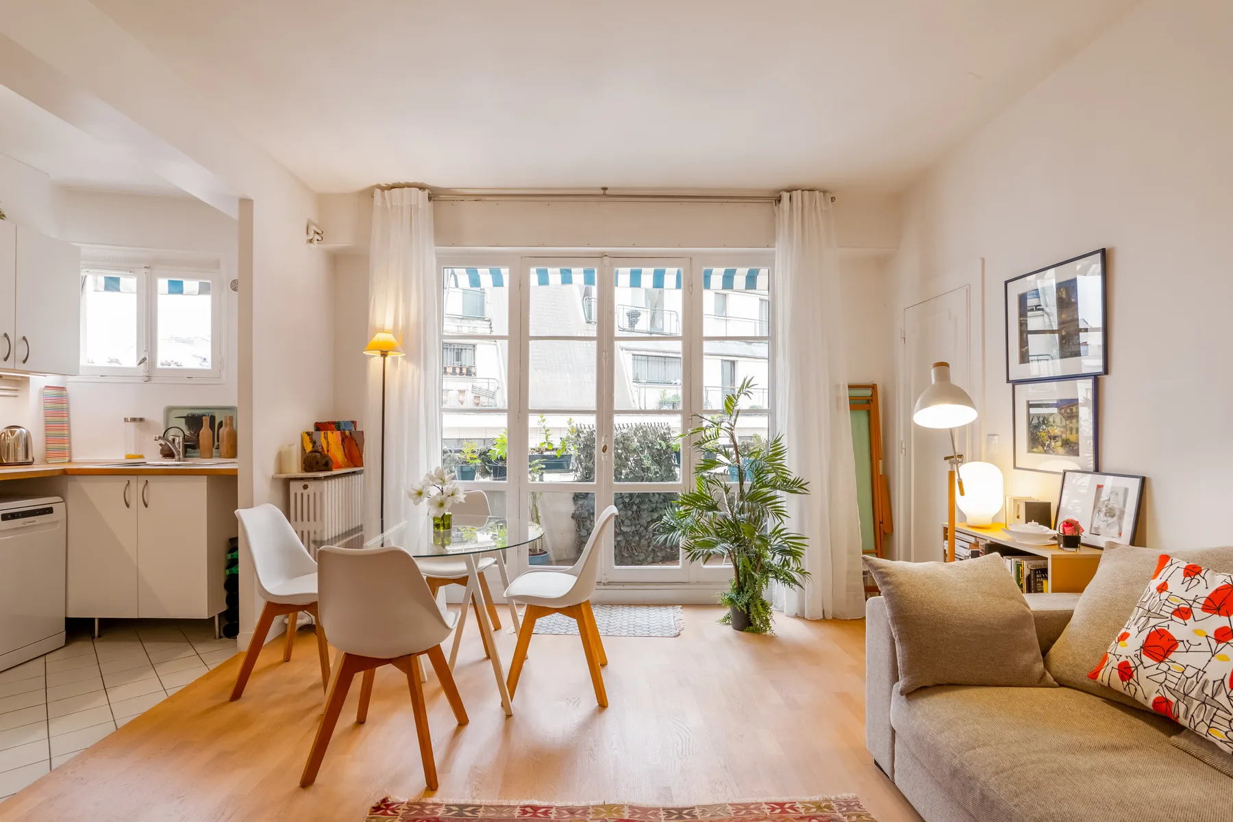 A bright living and dining area with large windows, seating, and a small kitchen.