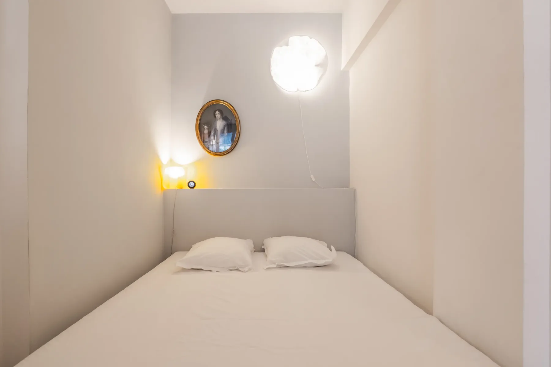 A narrow bedroom with a bed, two pillows, and a wall-mounted portrait.