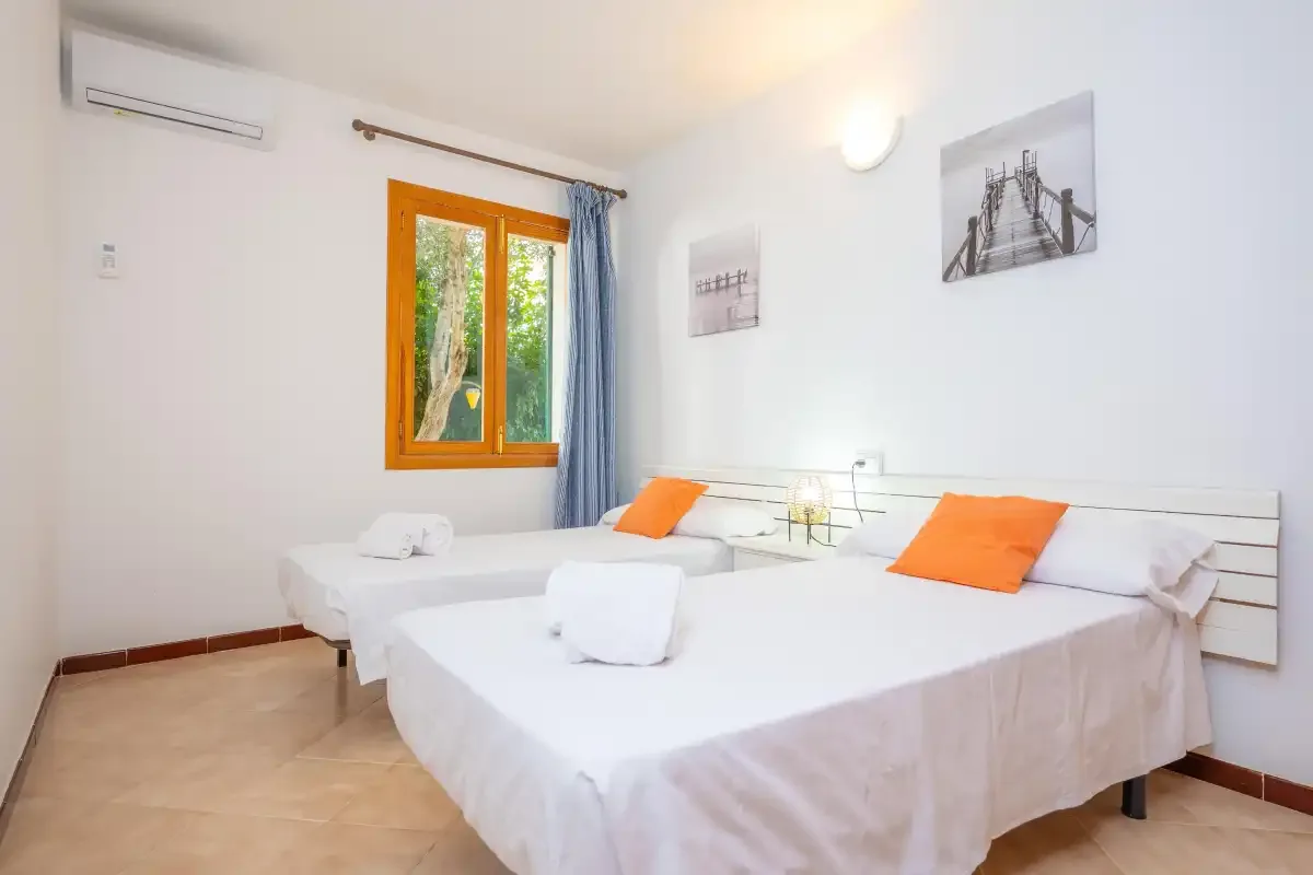 Un dormitorio luminoso con dos camas, cojines naranjas y una ventana.