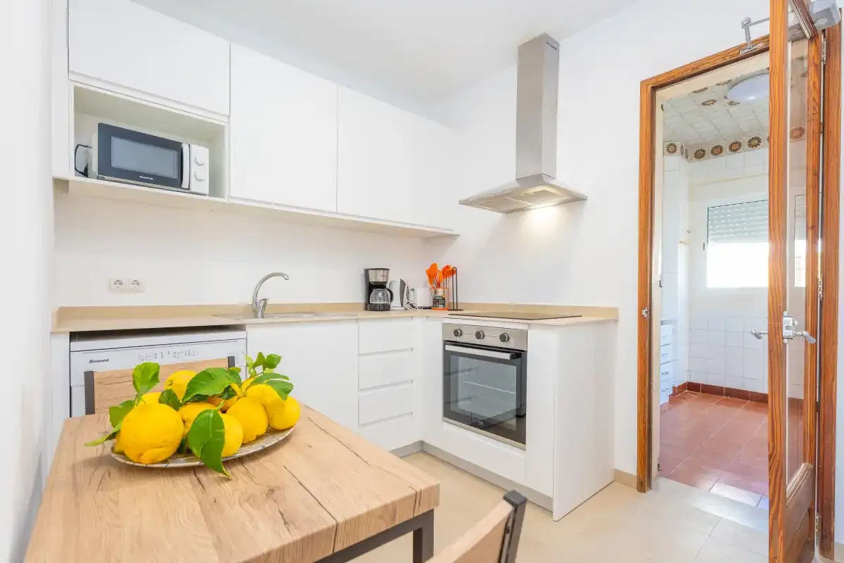 Una cocina luminosa con armarios blancos, una encimera y una mesa de madera.