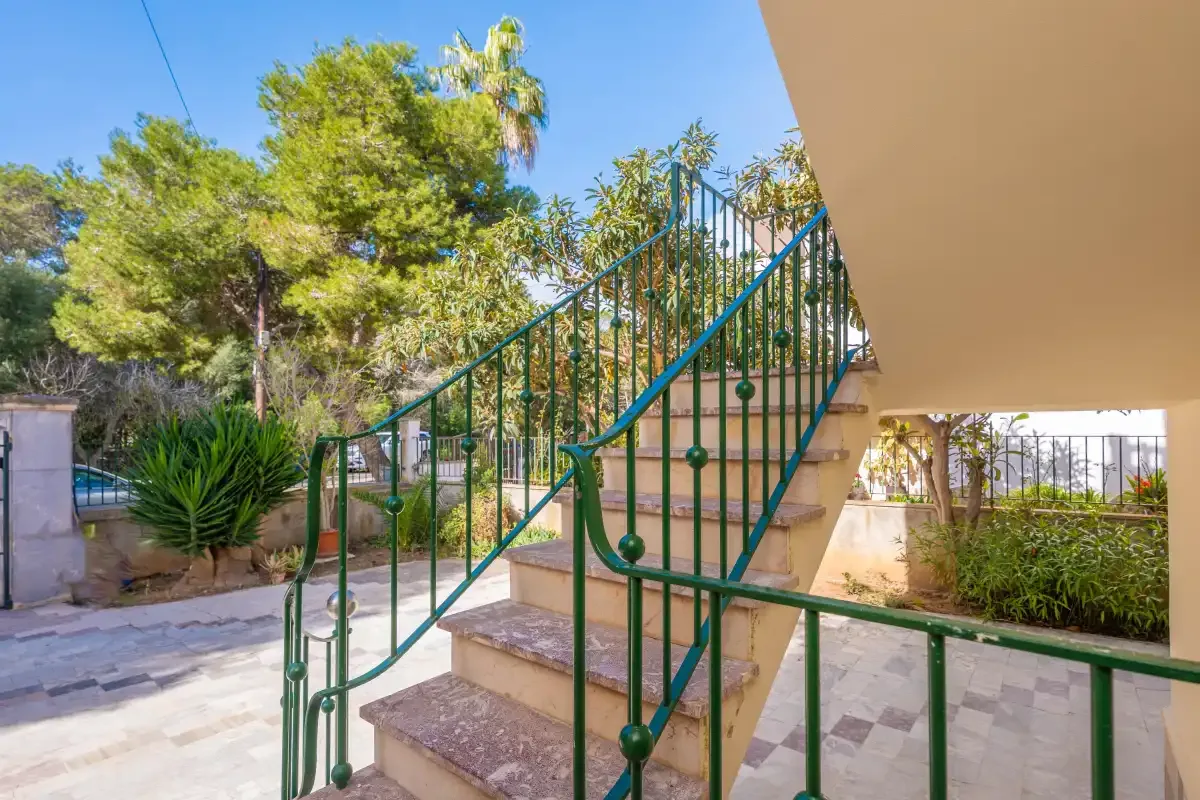Una escalera exterior con barandillas verdes está junto a un jardín ajardinado.