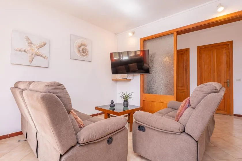 Sala de estar con asientos acolchados, una mesa de centro y un televisor en la pared.