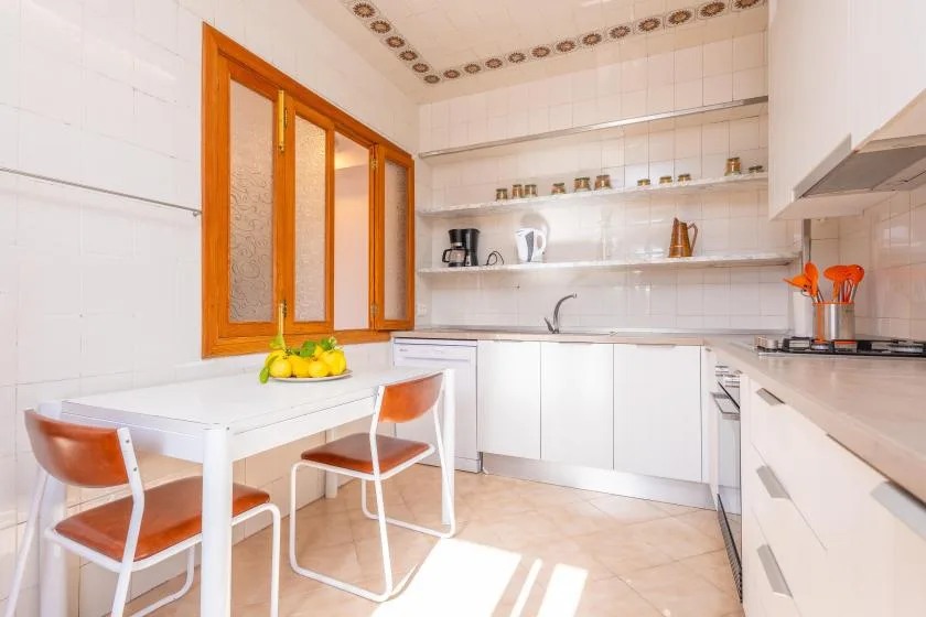 Una cocina luminosa con azulejos, armarios blancos, estantes abiertos y una pequeña mesa.