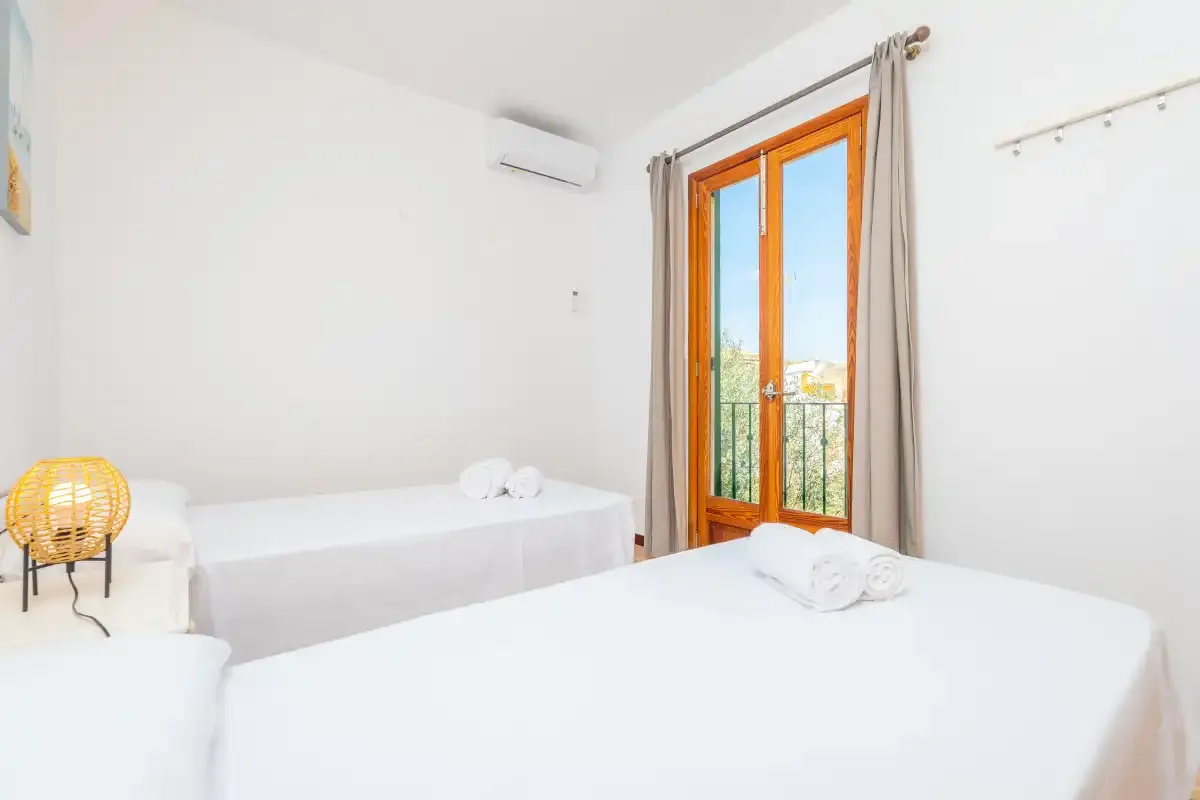 Un dormitorio luminoso con camas blancas y una puerta acristalada que da a un balcón.