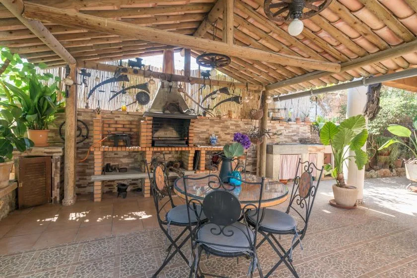 A covered outdoor dining area with a metal table and chairs beside a brick barbecue.