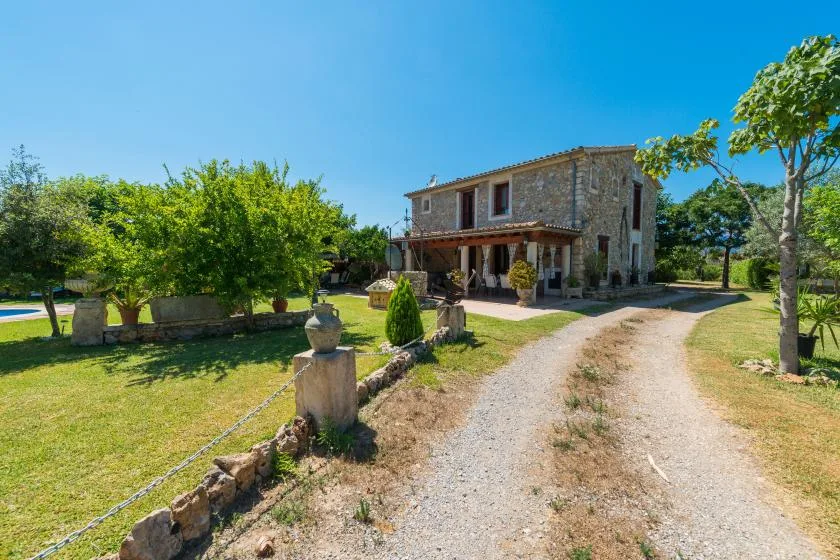 A stone house stands beside a gravel driveway in a sunny landscaped yard.