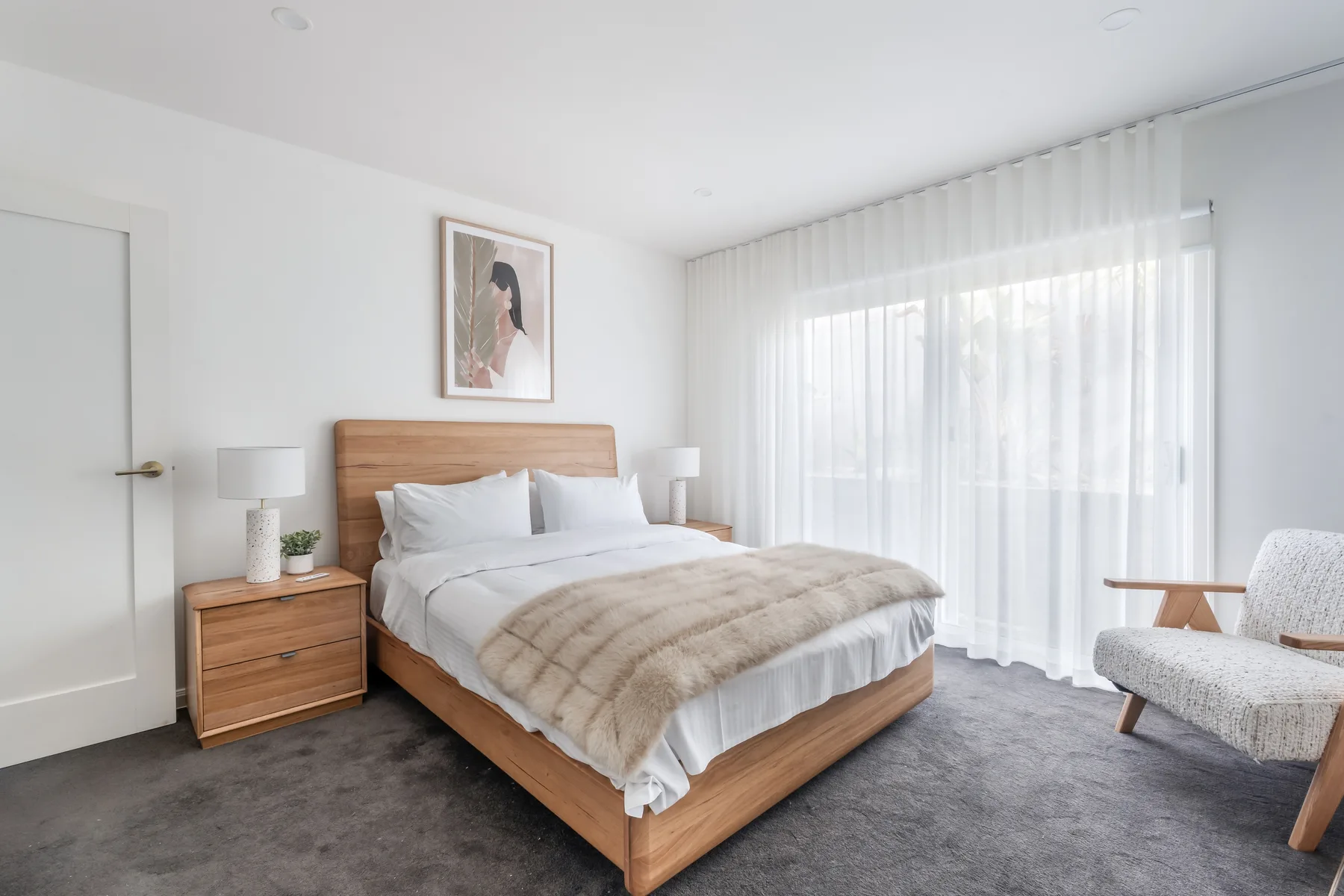 A bright bedroom with a neatly made bed beside a large curtained window.