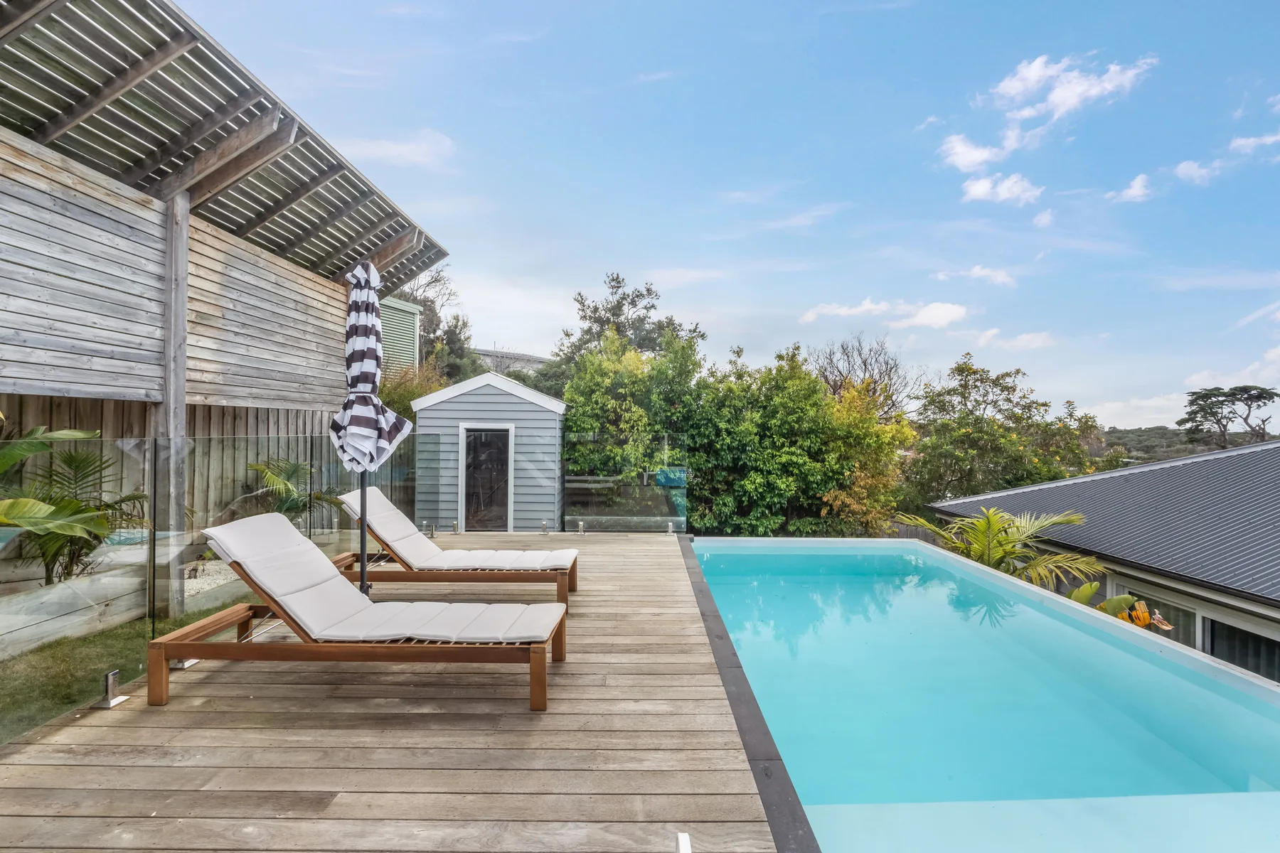 An outdoor swimming pool beside a wooden deck with lounge seating and surrounding greenery.