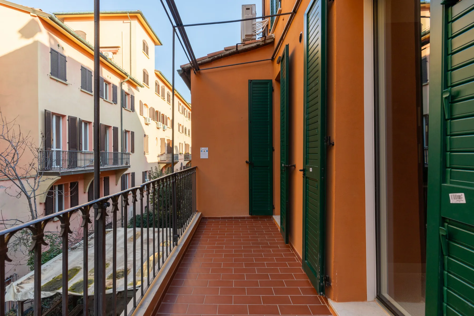 Una terraza exterior estrecha bordea un edificio naranja, con contraventanas verdes y una barandilla metálica.