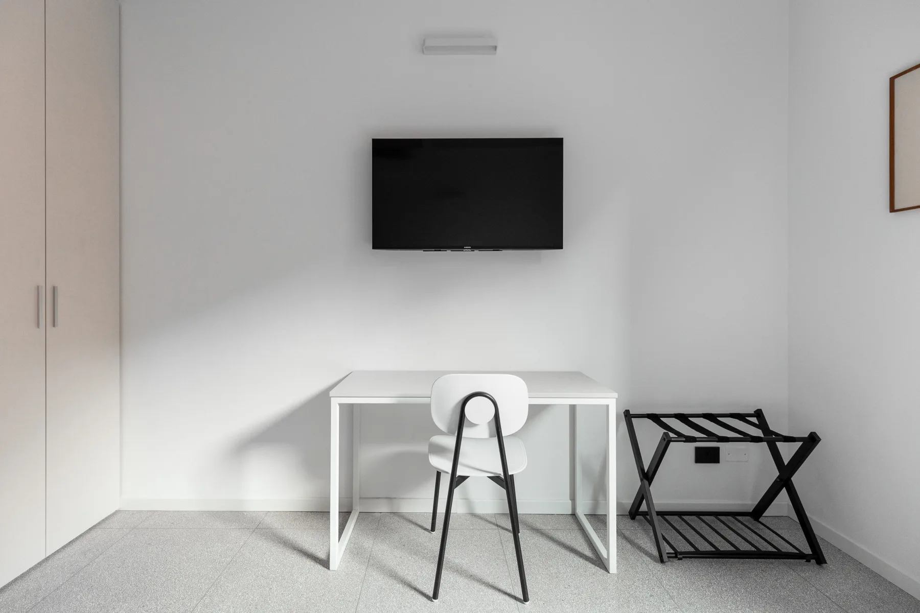 Una habitación luminosa con escritorio, silla, televisor en la pared y portaequipajes plegable.