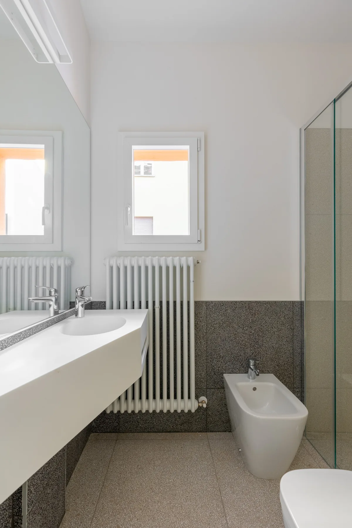 Un baño luminoso con una encimera de lavabo larga, un radiador y una mampara de ducha de vidrio.
