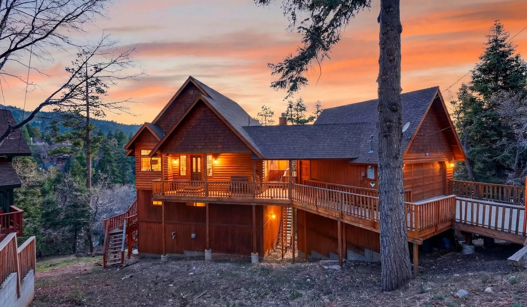 A wooden cabin with a wraparound deck glows beneath a colorful sunset sky.