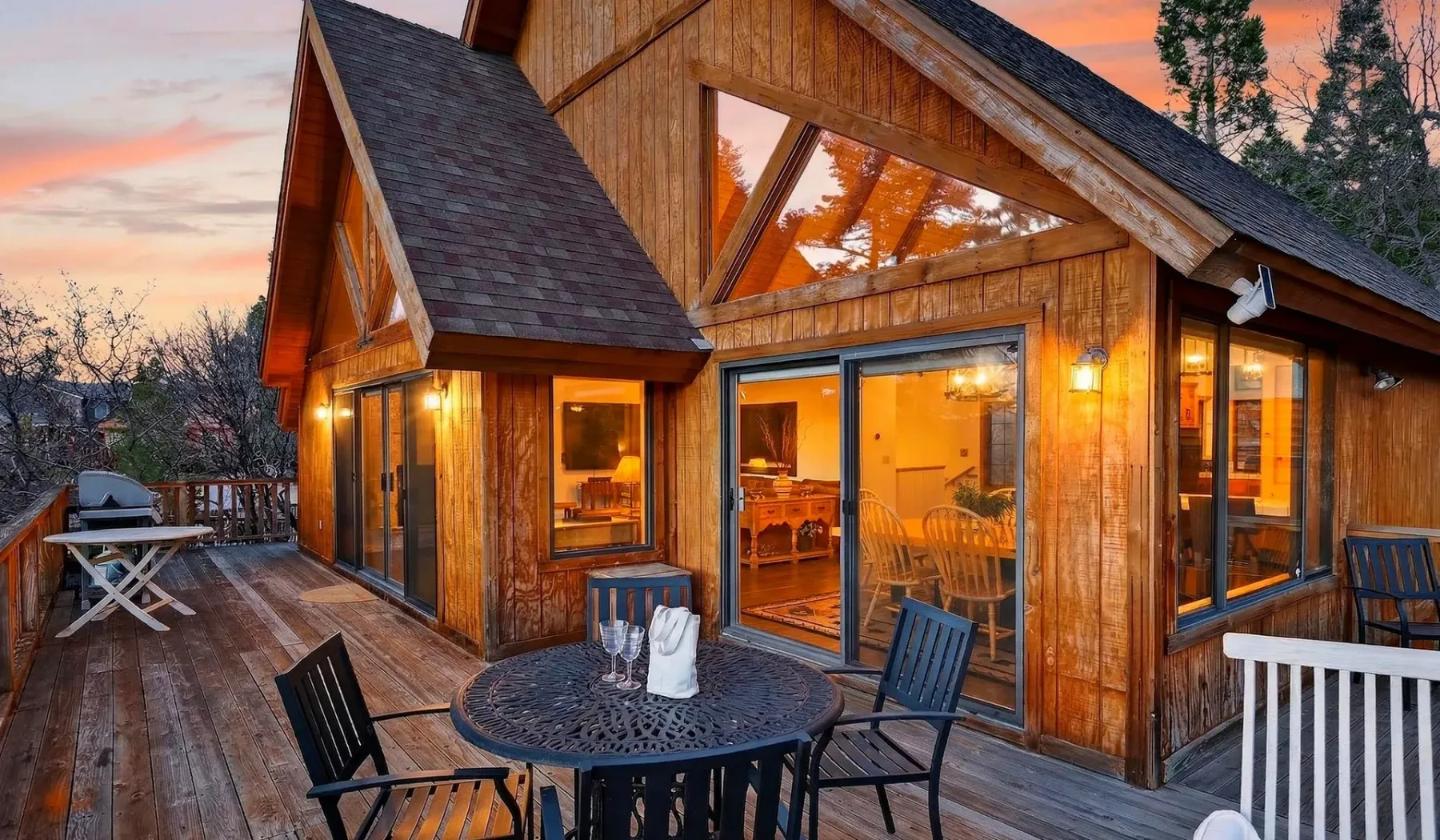 A wooden cabin with a lit interior and outdoor seating at dusk.
