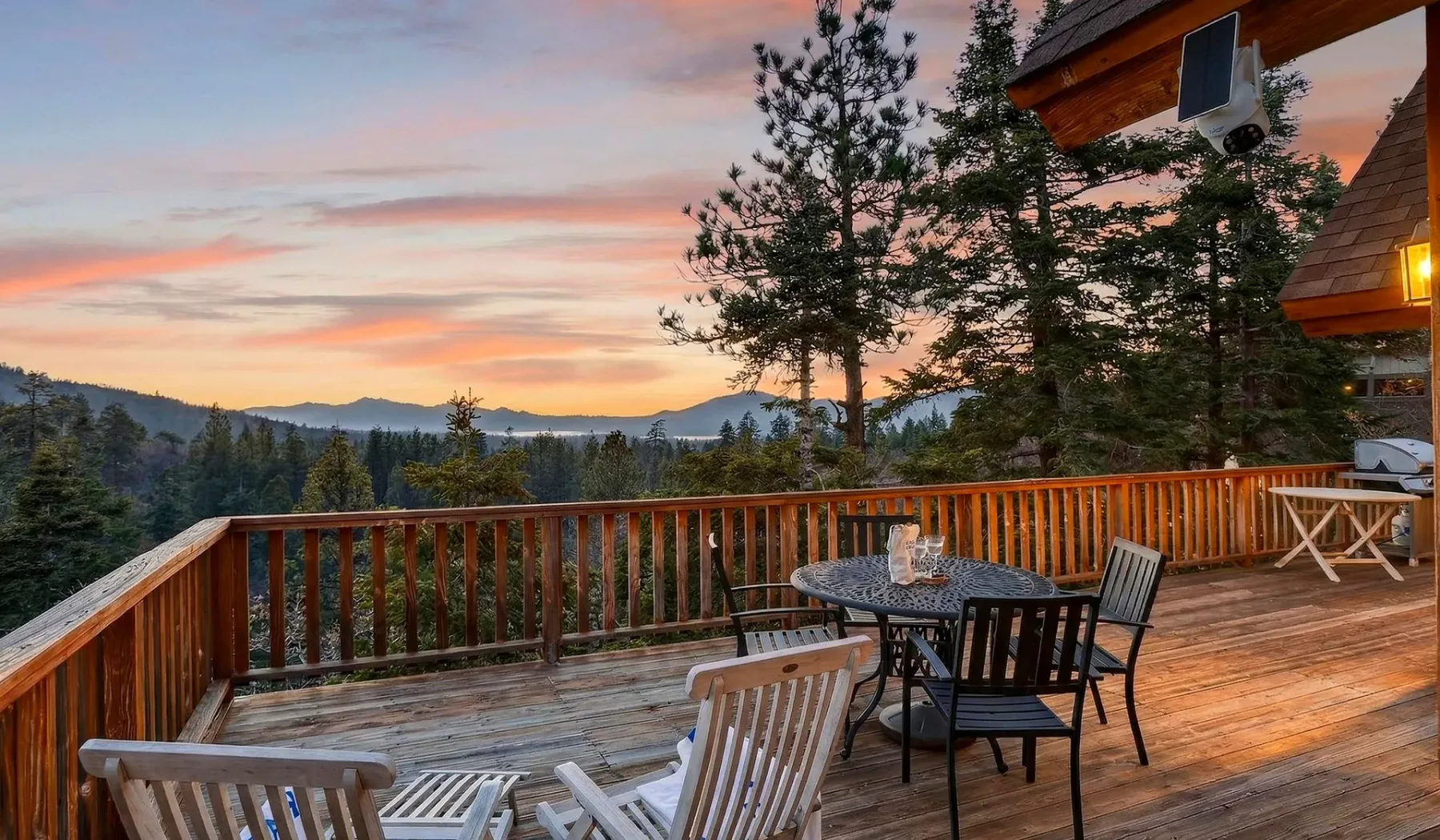 A wooden deck with outdoor seating overlooks a forested mountain view at sunset.