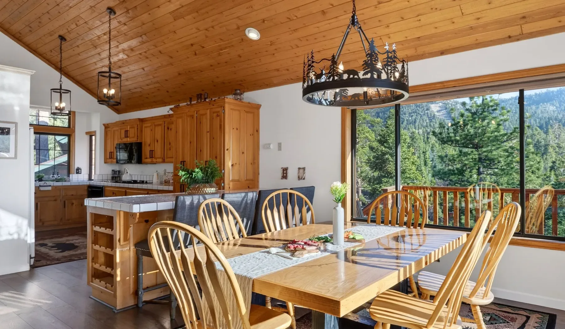 A dining area with a long wooden table beside large windows overlooking greenery.