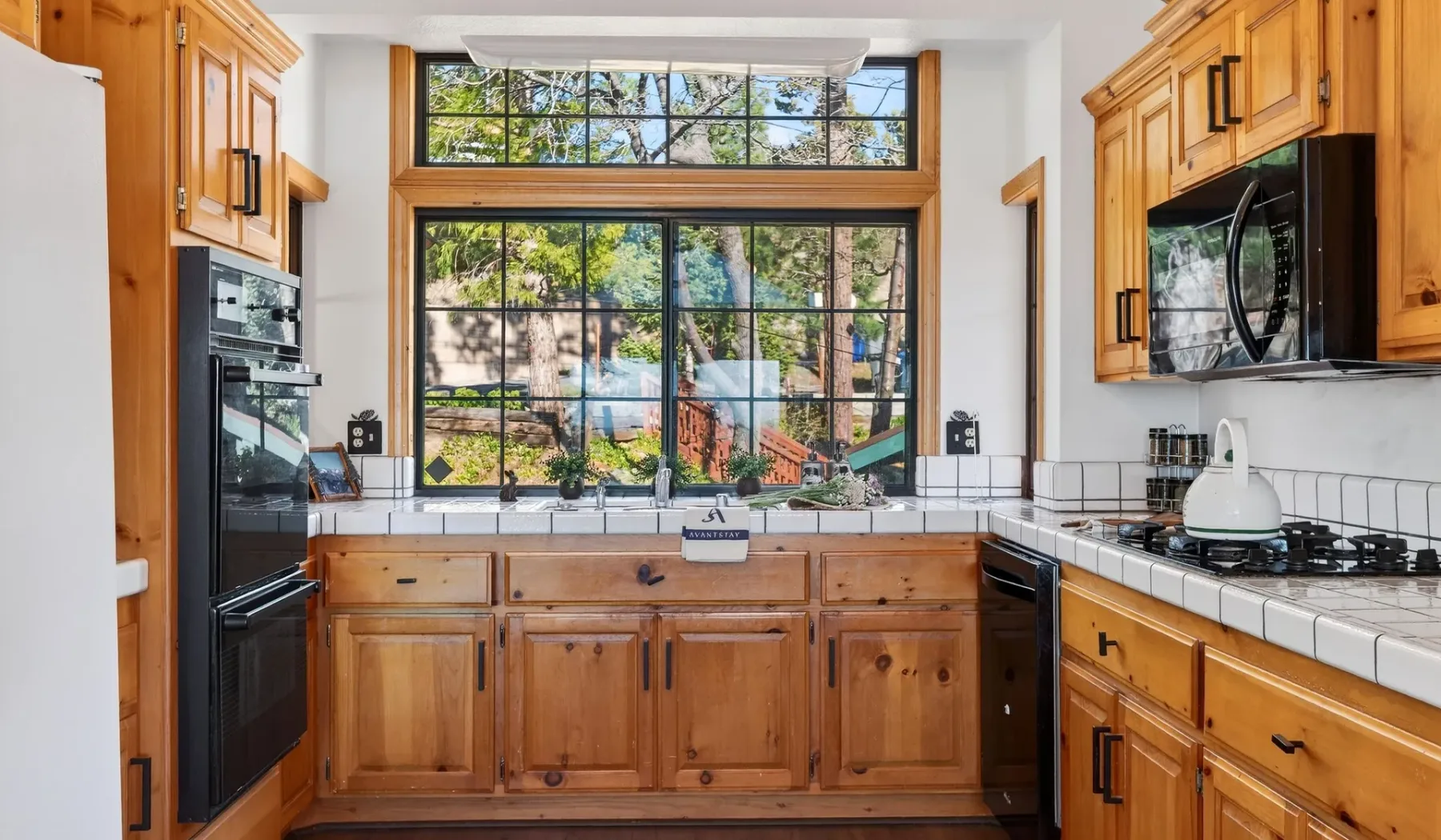 A bright kitchen with wooden cabinetry, dark appliances, and a large window.