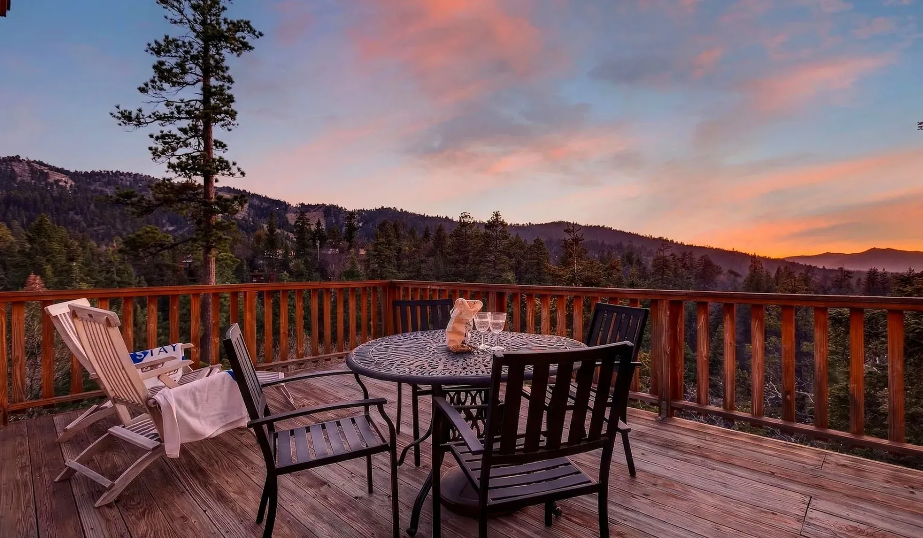 A wooden deck with outdoor seating overlooks wooded hills beneath a colorful sunset sky.