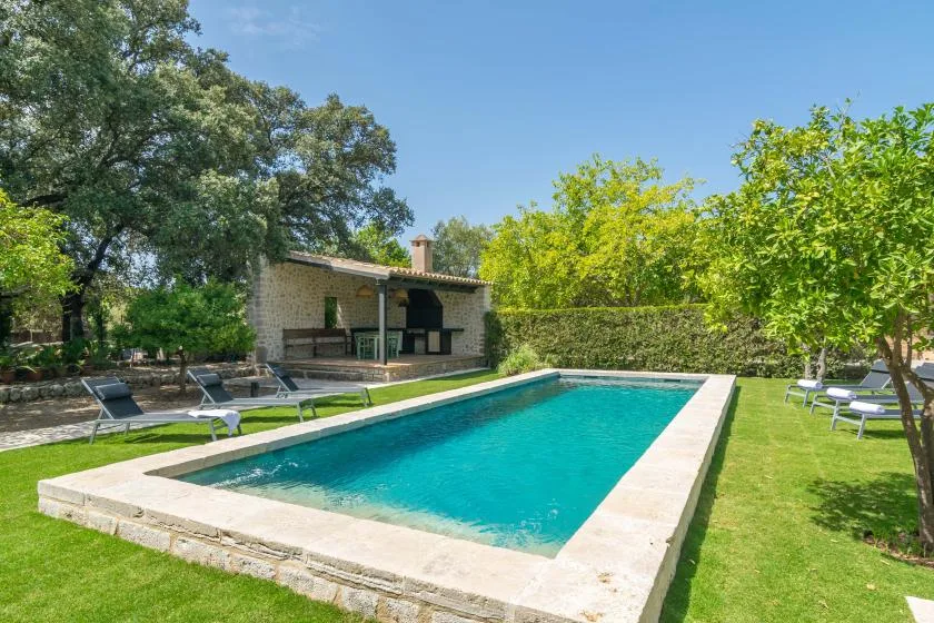 A rectangular swimming pool sits beside a shaded outdoor seating area in a green garden.