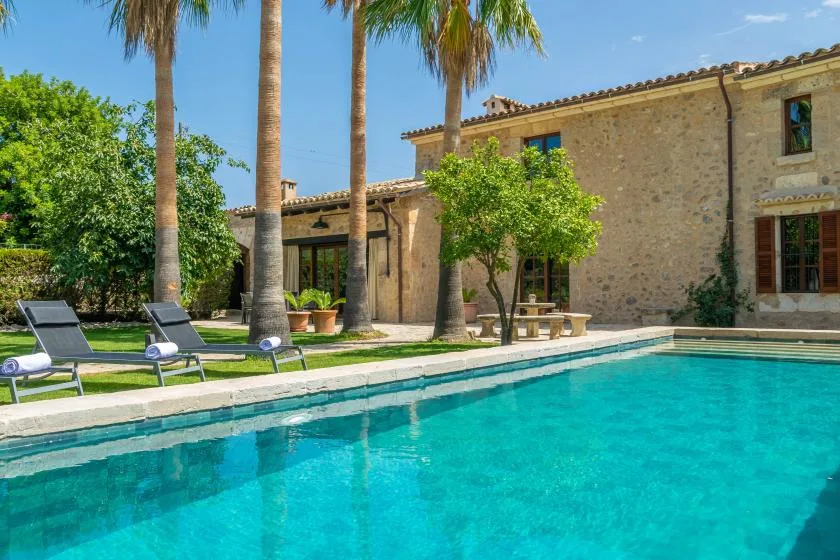 A swimming pool beside a stone building, with palm trees and lounge seating.