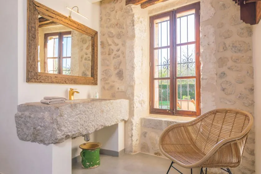 A stone-walled bathroom has a basin, tall window, and woven seating.