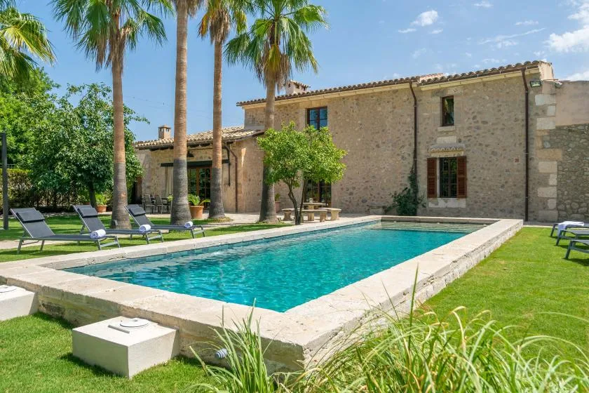 A swimming pool sits beside a stone building, with palm trees and lawn nearby.