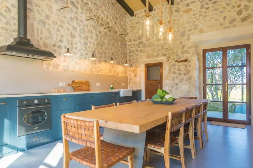 A rustic kitchen with blue cabinets, a large wooden dining table, and glass doors.