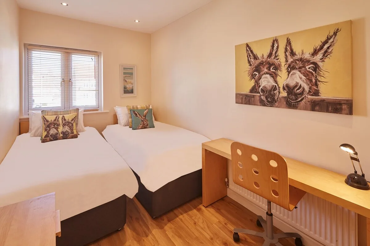 A bright bedroom with two beds, a window, and a wooden desk.