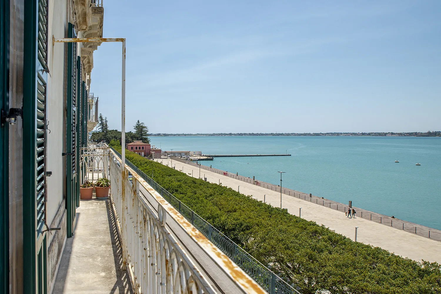A seaside balcony overlooks a waterfront promenade and calm blue water.