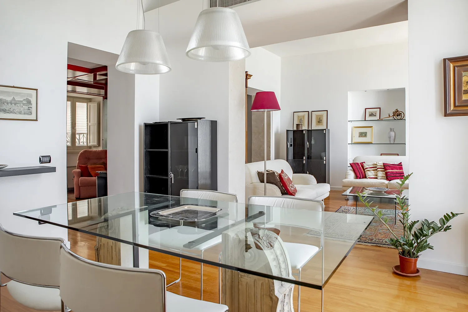 A bright living area has a glass dining table, seating, and shelving.