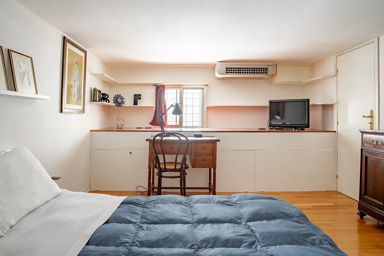 A bedroom with a bed in the foreground and a desk beneath the window.