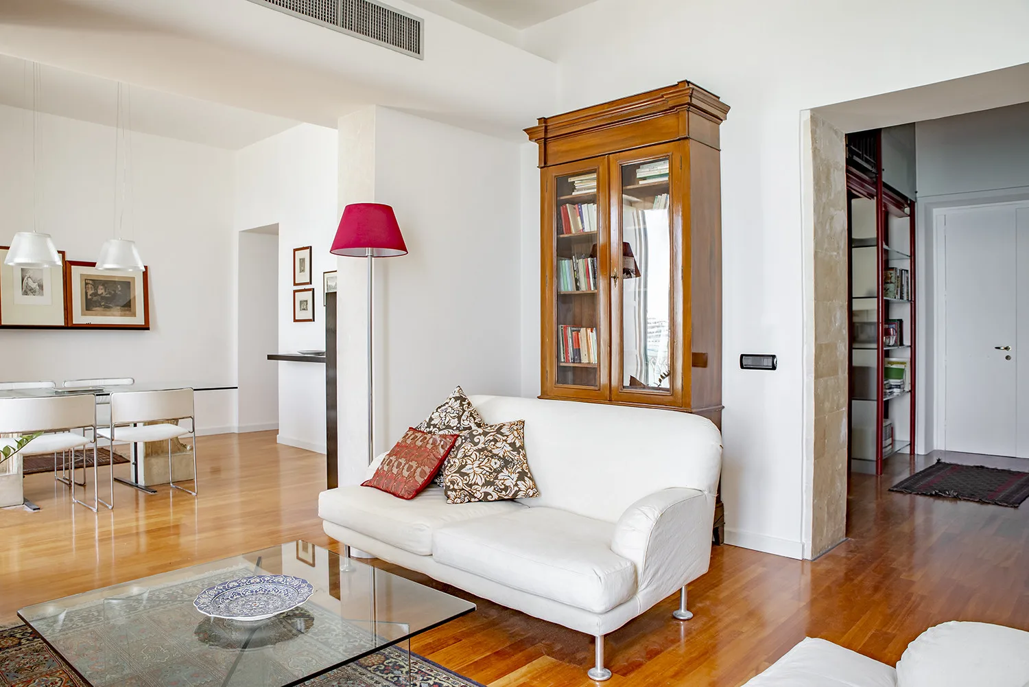 A bright living room with white seating, a glass coffee table, and a wooden display cabinet.