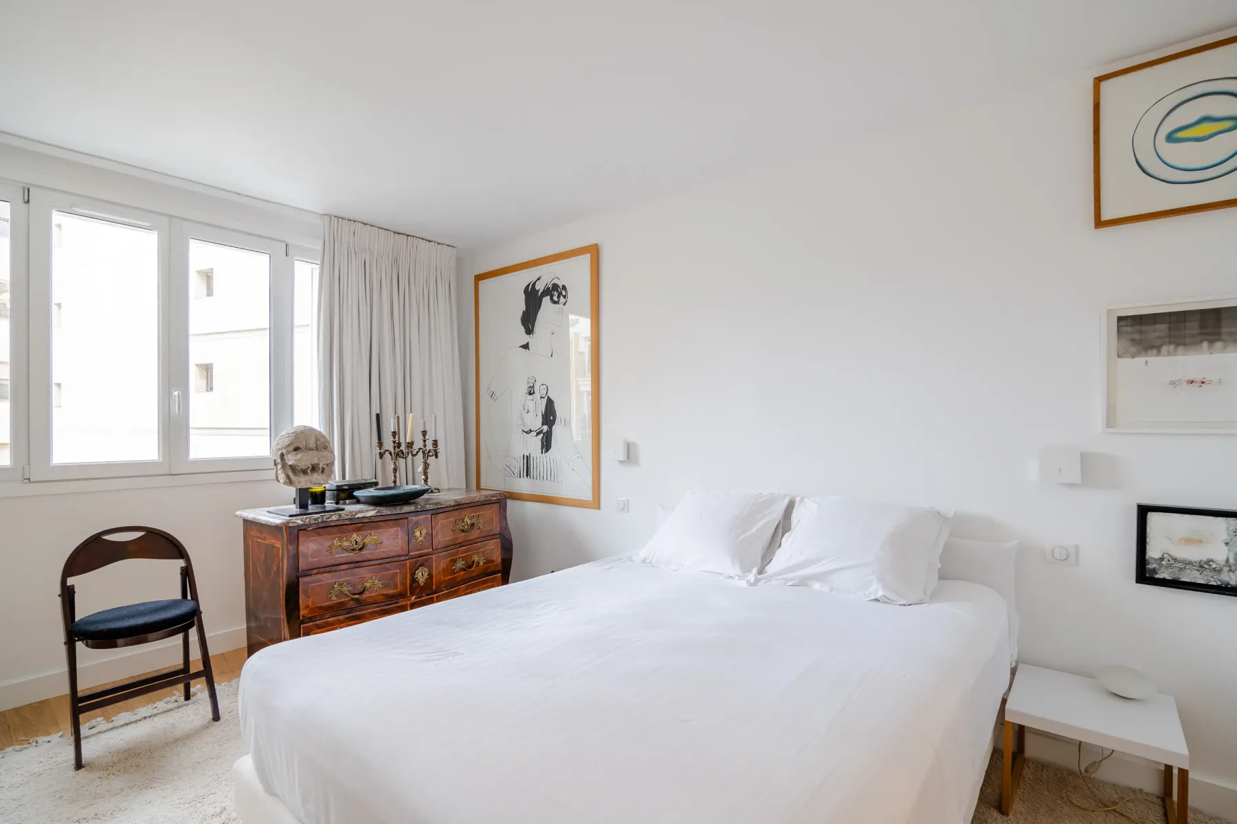 Un dormitorio luminoso con una cama blanca, cuadros enmarcados y una cómoda de madera.