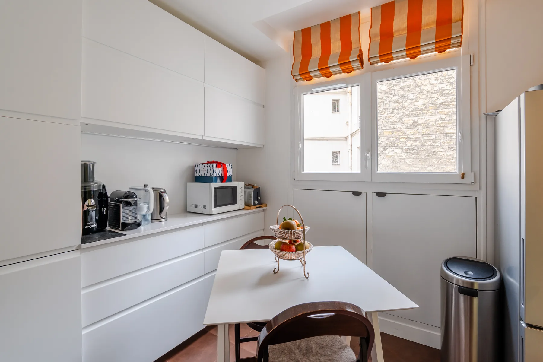 A bright kitchen with white cabinets, a dining table, and an orange-striped window shade.