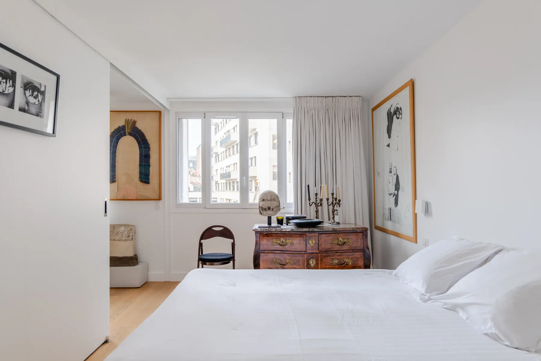Un dormitorio luminoso con una cama blanca, obras enmarcadas y una cómoda de madera.
