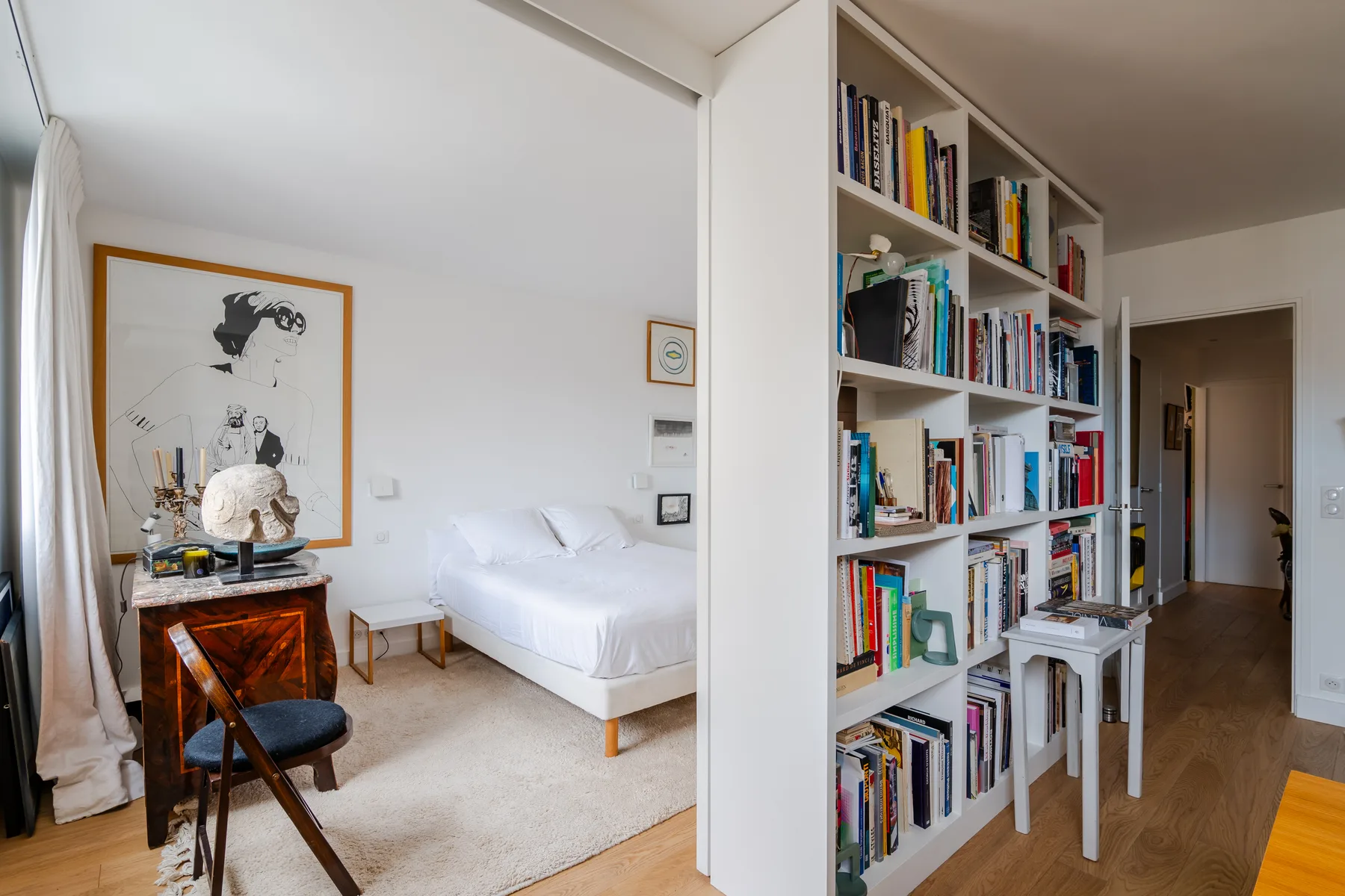 Un dormitorio luminoso con una cama blanca y una estantería alta junto a la entrada.