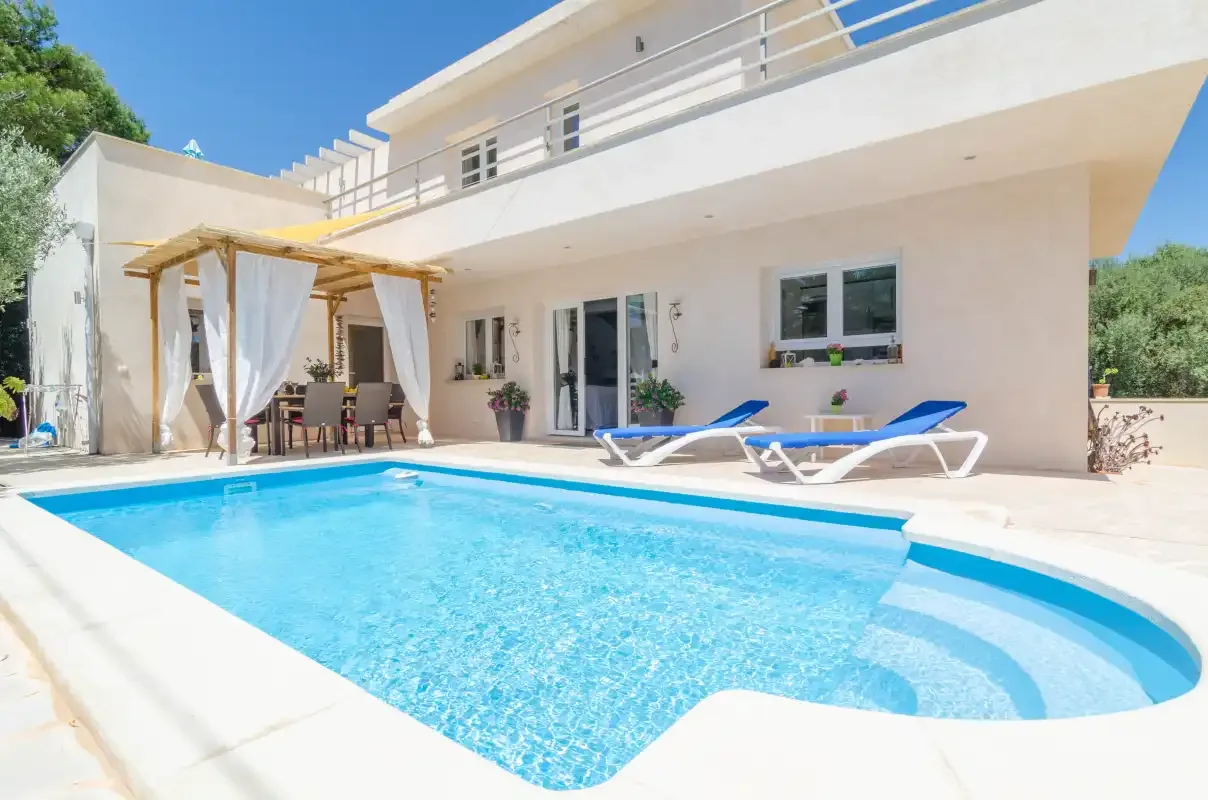 A swimming pool sits beside a modern white house with a shaded patio.